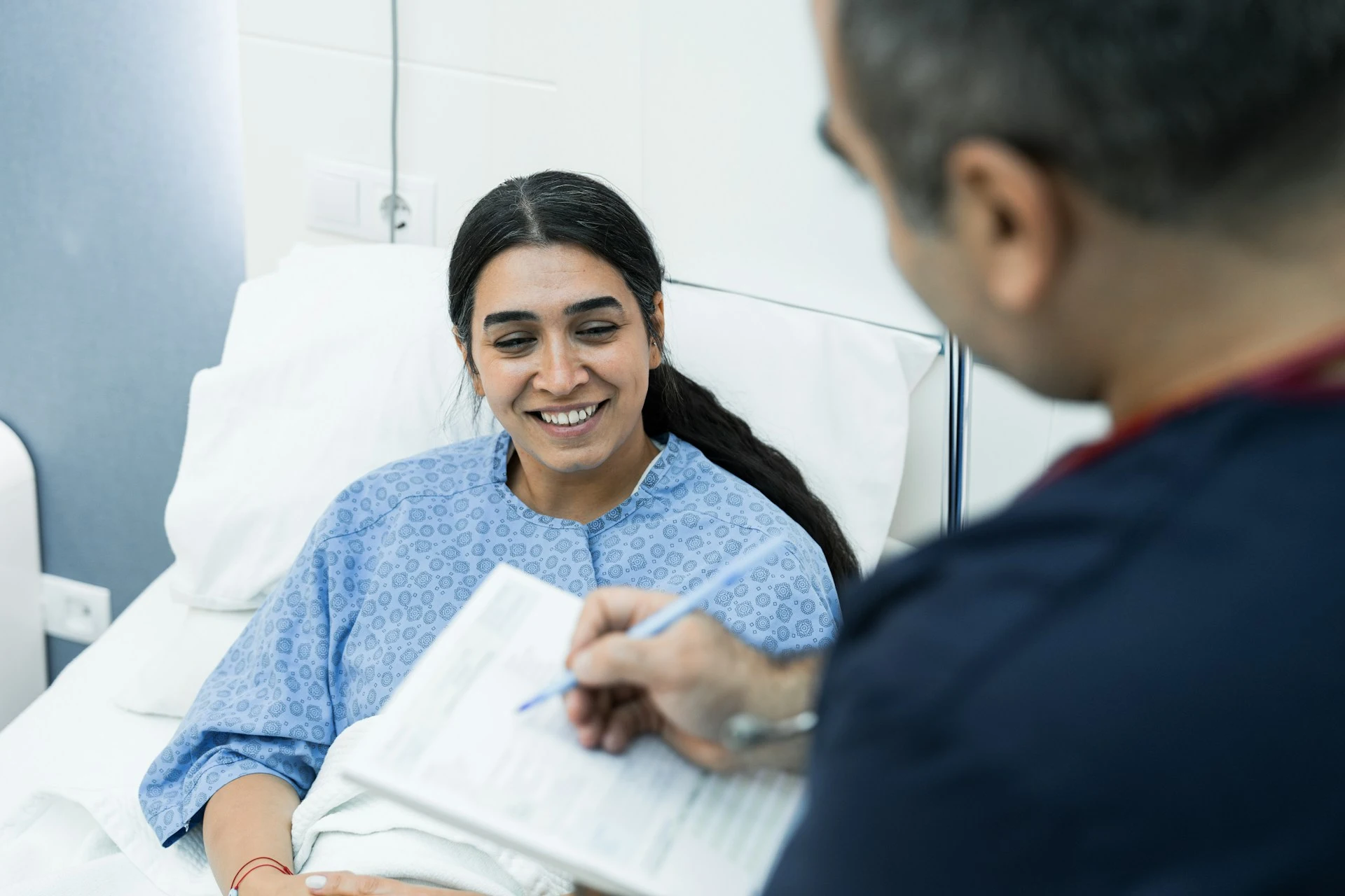A smiling patient in a hospital gown speaking with a healthcare worker filling out paperwork.